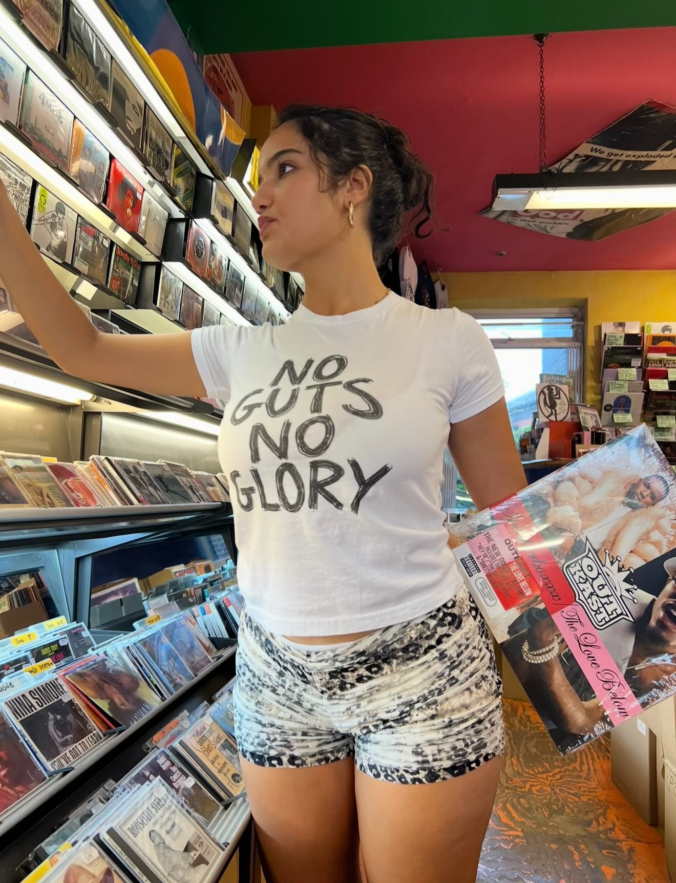 A woman standing in a record store aisle wearing a white graphic t-shirt that reads 'NO GUTS NO GLORY' and patterned shorts, holding a vinyl record.